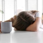 A tired woman puts her head down on her counter while holding a cup of coffee.