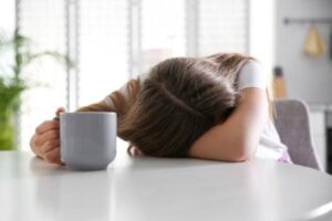 A tired woman puts her head down on her counter while holding a cup of coffee.